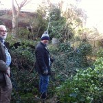 Two committee members infront of an overgrown fence, before work started to plant the area
